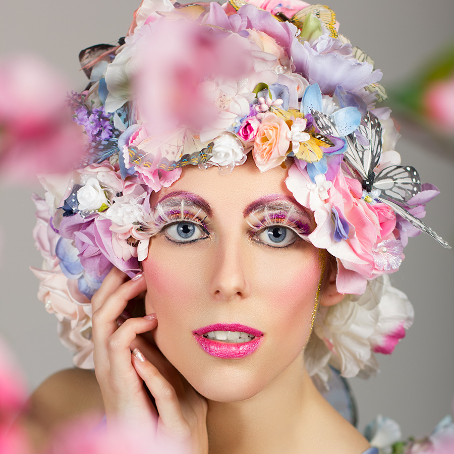 Through blurry pink cherry blossoms, a woman's face can be seen with fantastical sunrise-coloured makeup, including white feather eyelashes. Instead of hair, her head is covered in a riot of colourful silk flowers and feather butterflies.