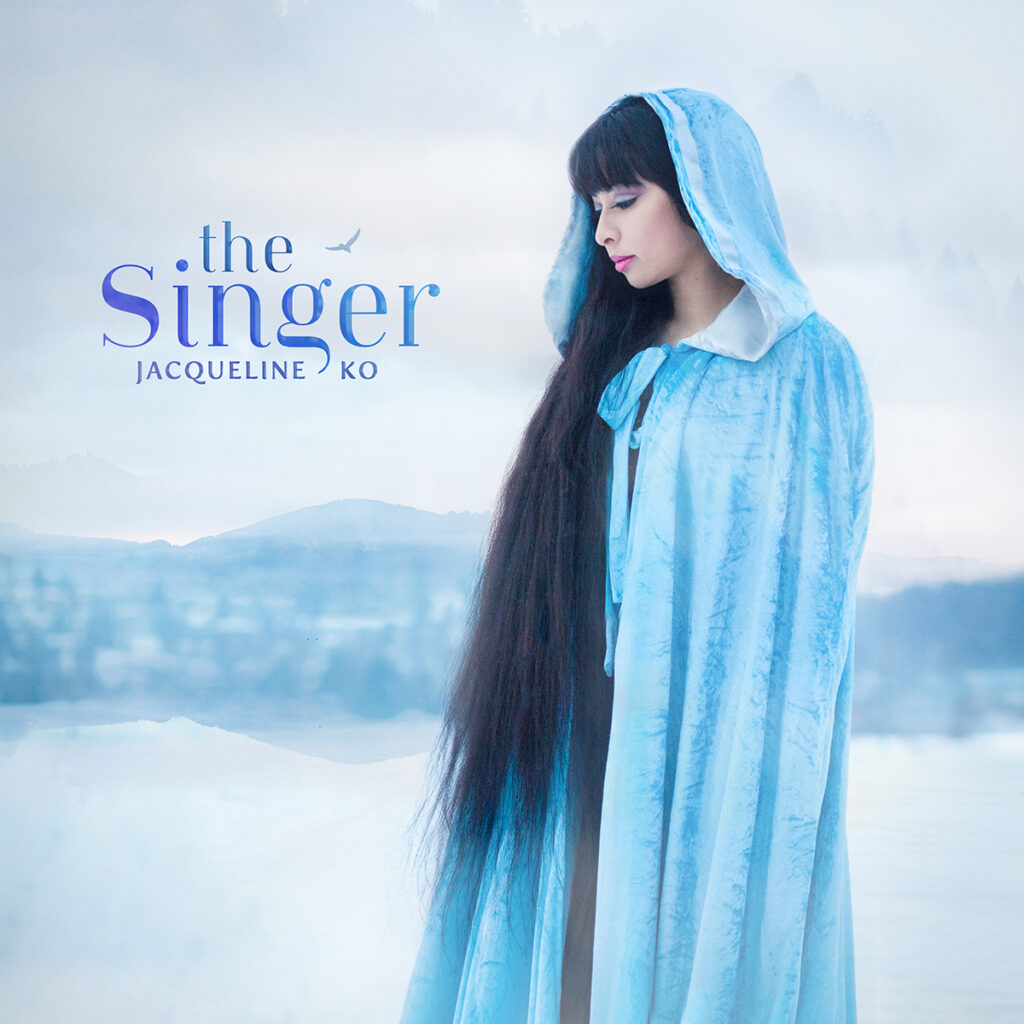 Against a misty blue background of mountains and a lake, an Asian woman with extremely long black hair stands in profile, wearing an ice blue velvet cloak and a serene expression. Blue text reads, The Singer. Jacqueline Ko.