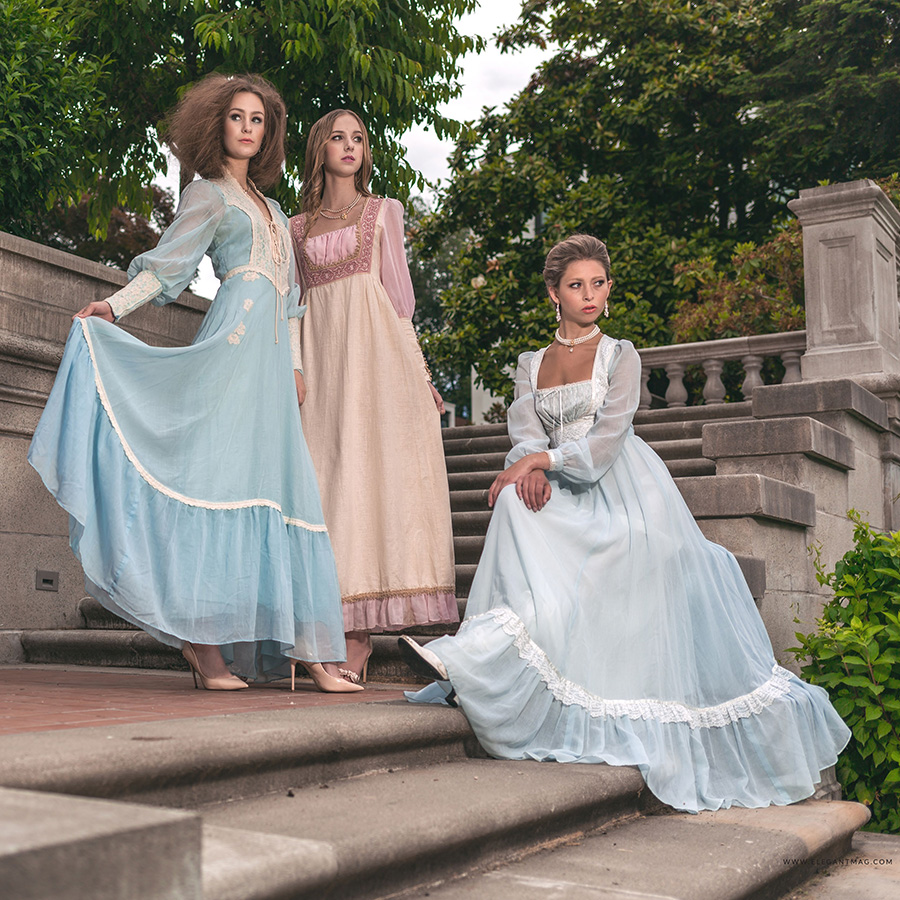 Three slender white women with blonde hair are standing and sitting on an elegant staircase outdoors, amid a garden. They are wearing delicate muslin dresses in pale blue, pink and cream, trimmed in lace and delicate embroidery details, with pearl necklaces and earrings. Their sweeping skirts drape down the stone steps and blow in the wind.