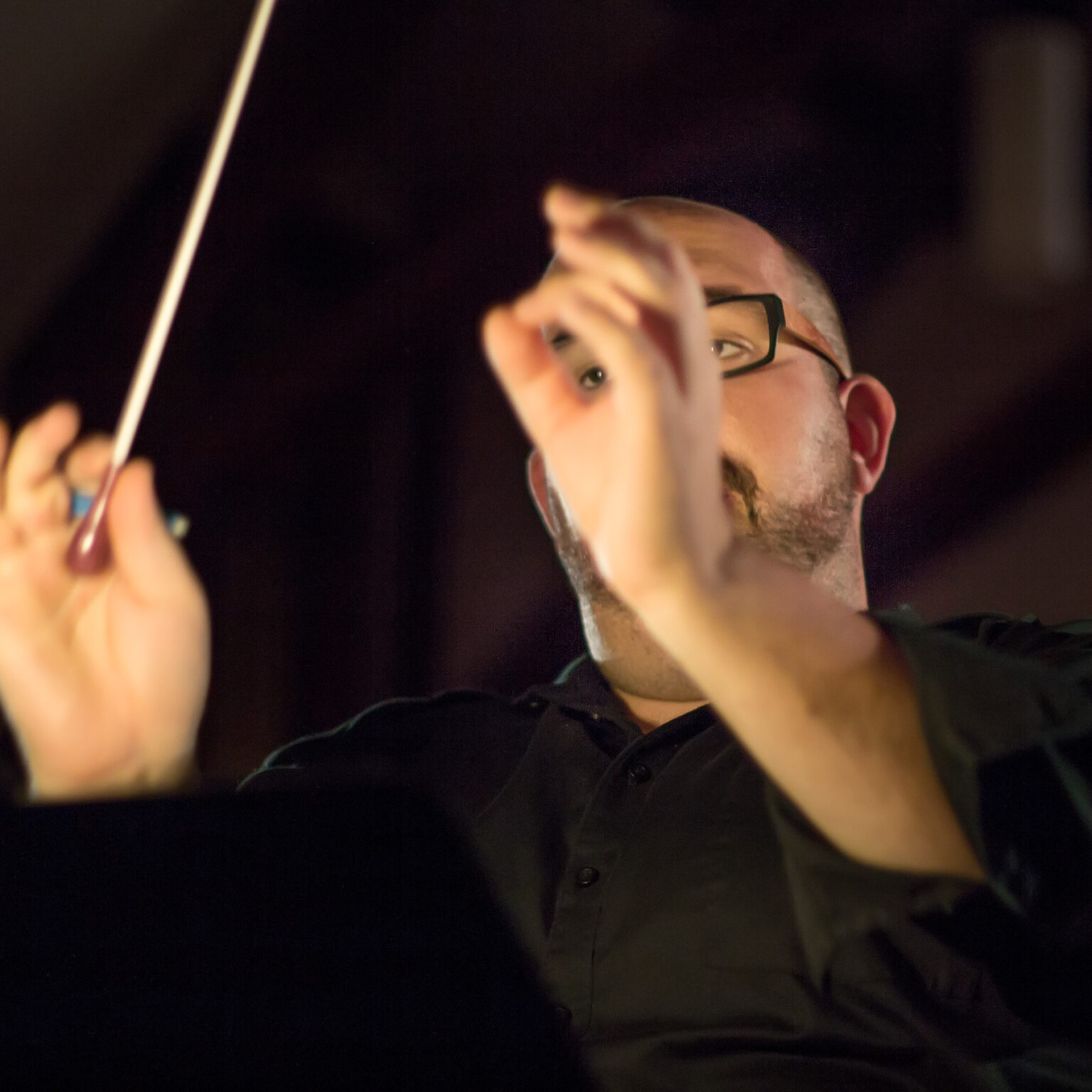 A white man with a beard and glasses wearing a black shirt is conducting in a shadowy theatre. He has his hands raised, with a baton in one hand and the fingers of the other hand cupped in such a way that the viewer can glimpse one of his eyes through his fingers.