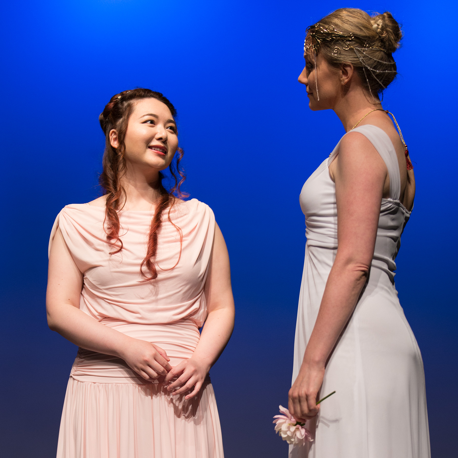 Onstage, two performers in pale Grecian gowns stand against a vibrant blue backdrop. On the left, a Chinese Canadian woman with long auburn hair is clad in pale pink, and smiles up at a taller white woman with honey coloured hair clad in white, wearing a filigreed golden crown and holding a large pink blossom in her hand.