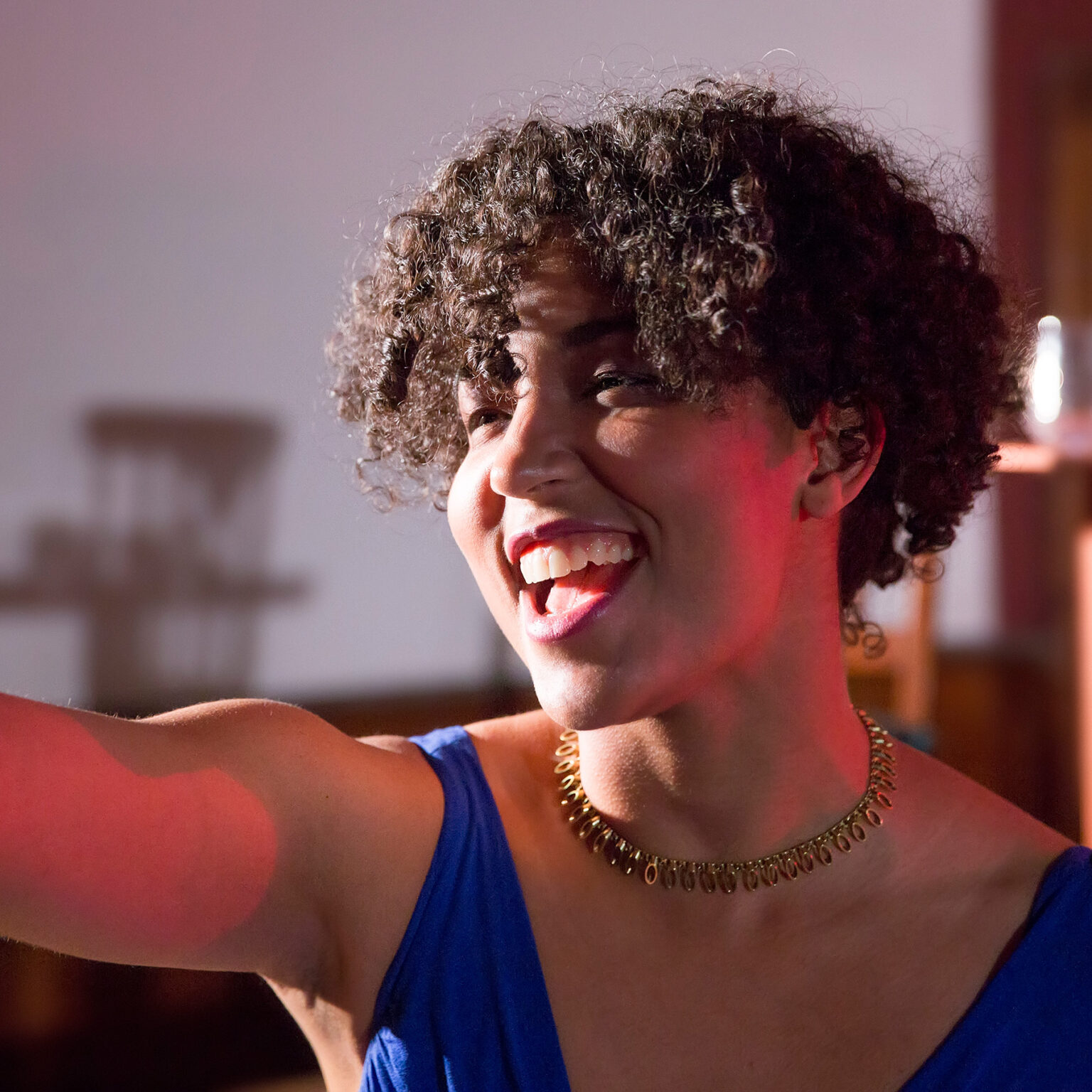 A Black non-binary person with natural hair is onstage, wearing a blue top and gold necklace, bathed in red light. They have one arm outstretched, and a joyous smile on their face.