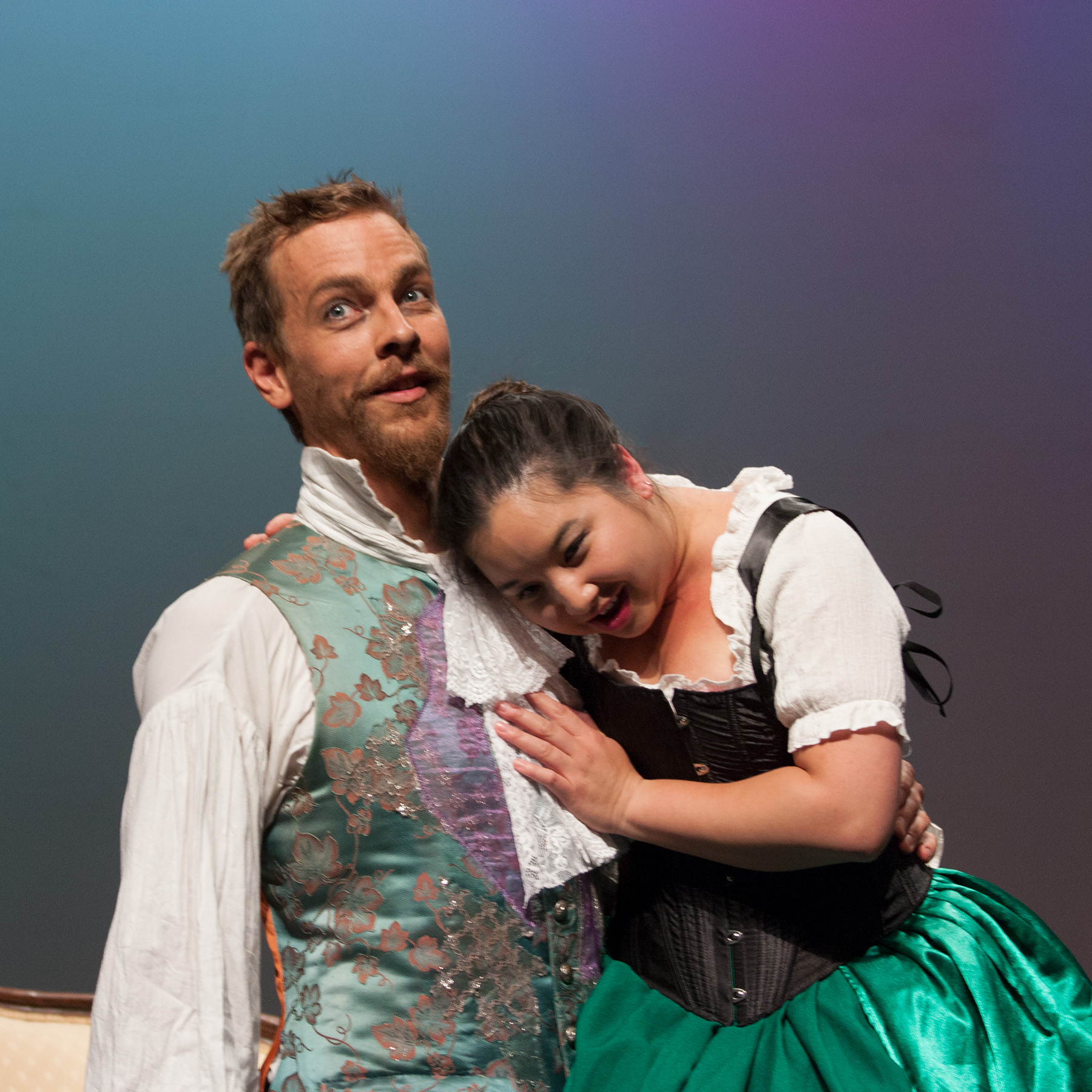 Onstage, a tall white man in a mint green 18th century waistcoat looks out towards the audience with an expression of incredulous joy, as on his lap, a pretty Chinese Canadian woman in a black corset and green skirt nestles her head against his chest, listening to his heartbeat with a mischievous expression.