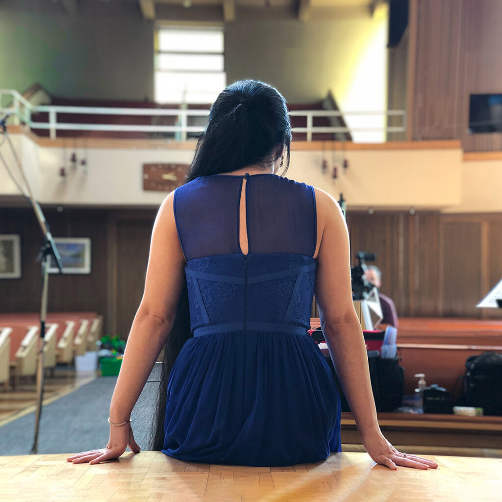 Back view of a young woman seated onstage, wearing a blue gown, with her very long black hair trailing on the floor. Around her is a light-filled church sanctuary. In the blurry distance can be seen various microphones on tall stands, as well as the small figure of a cameraman with a video camera.