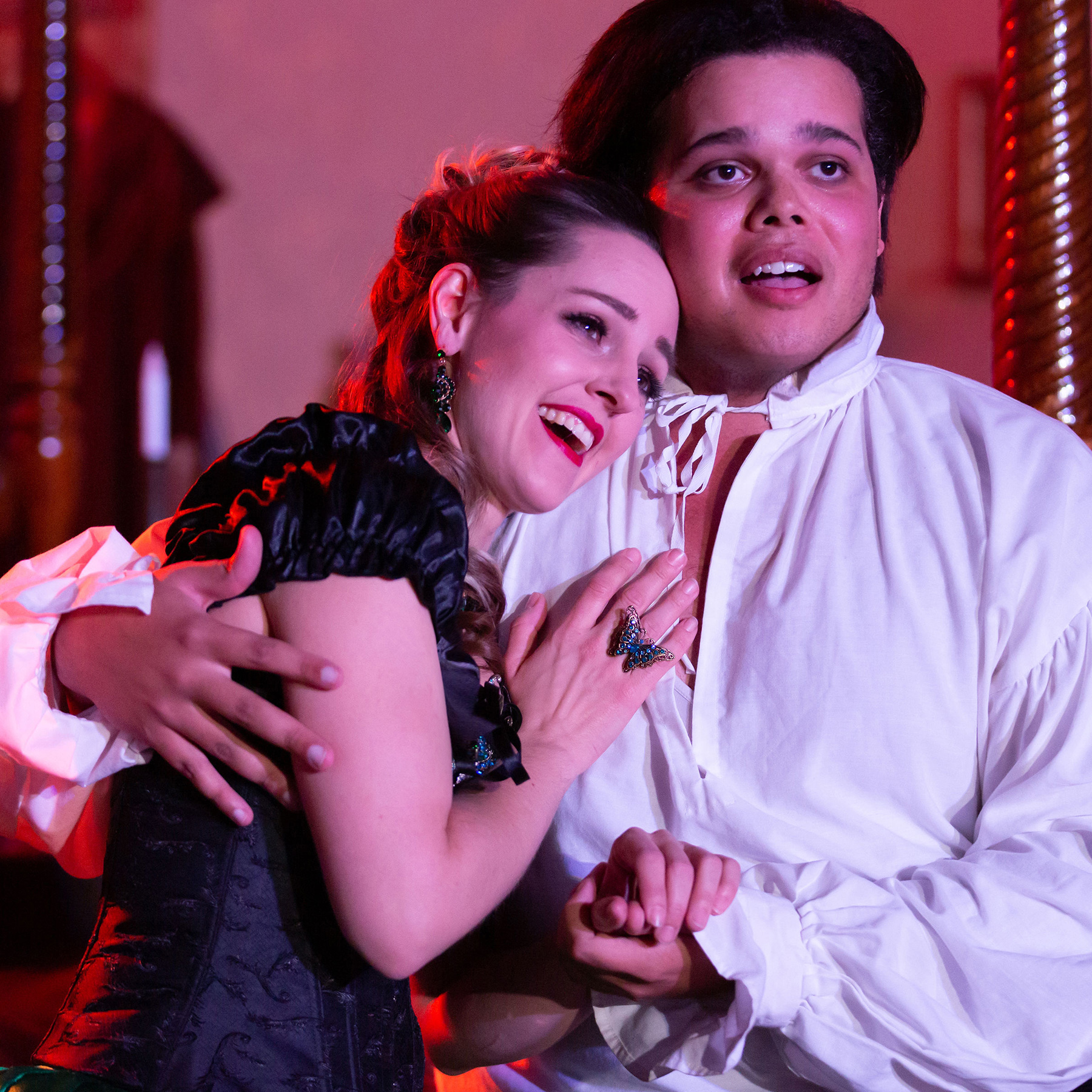 Bathed in rosy light, two performers have their arms around each other, gazing into the distance with dreamy smiles - on the left, a beautiful blonde woman wearing a ruffled black Victorian top and crystal jewelry, and on the right, a young Black man wearing a white poet shirt.