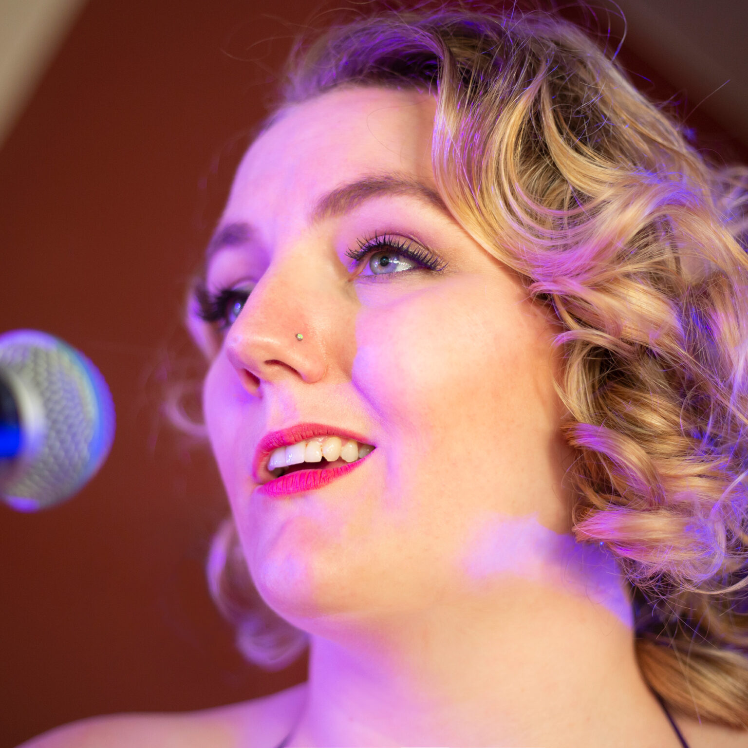 A closeup headshot of a white nonbinary person with blond ringlets, long lashes and a small crystal nose stud, singing into a microphone onstage