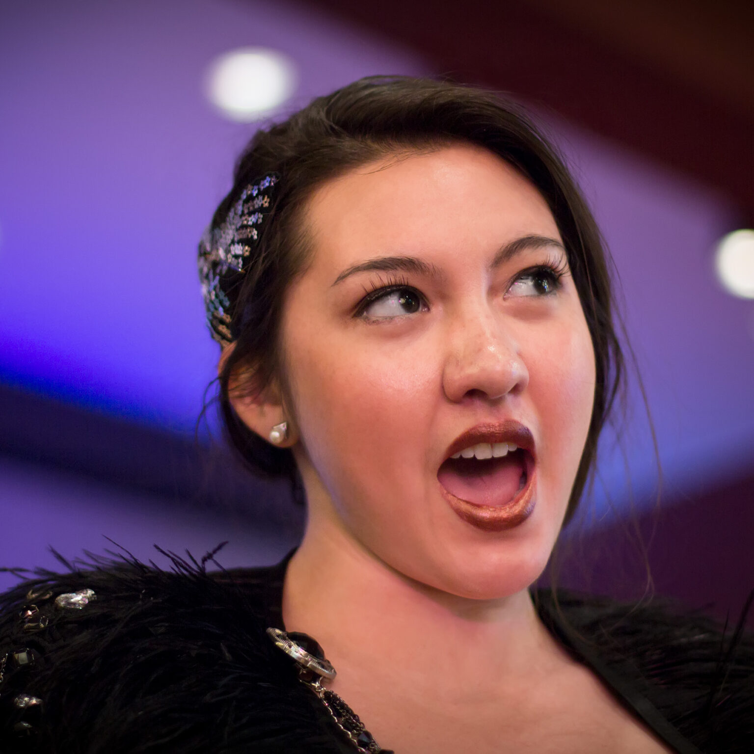 A brunette woman of mixed racial heritage is performing onstage with a sly expression, lit by blue and purple light. She wears deep red lipstick, a feathery black cape and a glittering silver hair clip shaped like a butterfly.