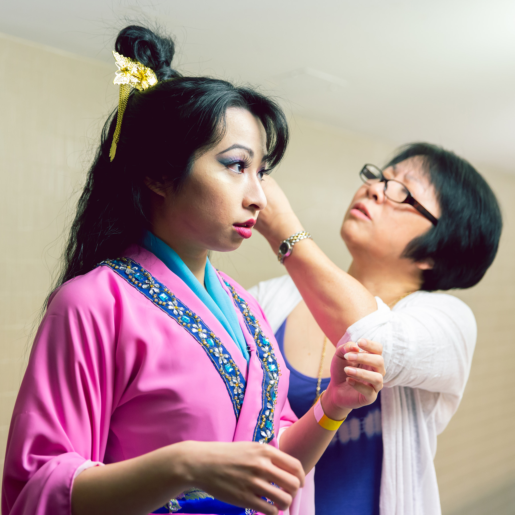 A young Asian Canadian woman dressed in a traditional Chinese outfit of pink and blue is standing still in the foreground. Behind her, a middle-aged Asian Canadian woman in casual clothing has her hands uplifted, carefully working on the first woman's intricate traditional updo, inserting golden filigree pins into it.