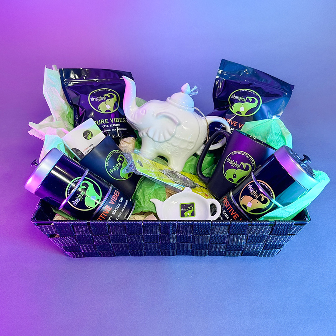 Against a blue and purple background, a black woven basket is full of Chai Ghai organic teas, mugs, and tea accessories including a white elephant-shaped teapot