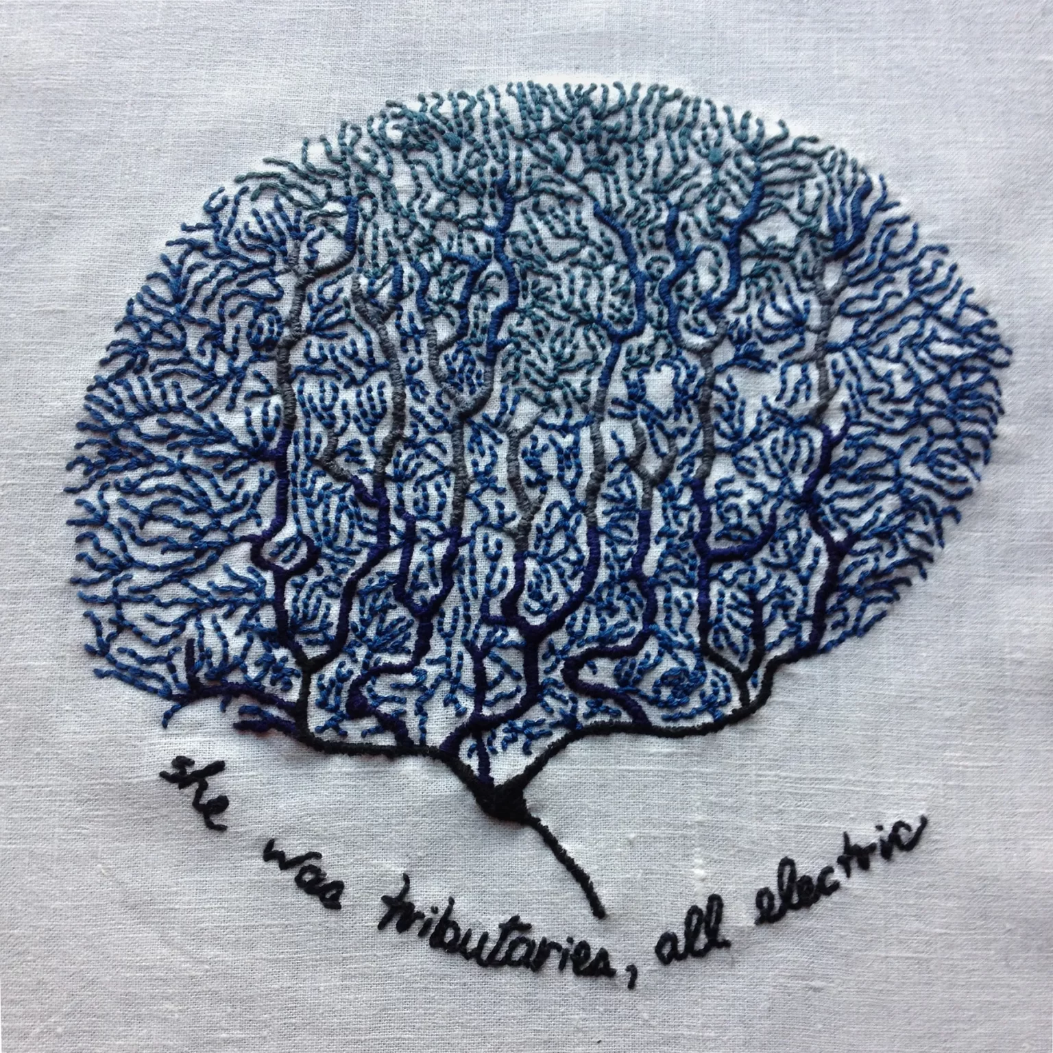 An embroidery on bone white linen. There are many coral-like neurons in the vague shape of a brain embroidered in shades of blue. The bottom section is navy blue and transitions to a lighter blue at the top. The text at the bottom reads: she was tributaries, all electric.