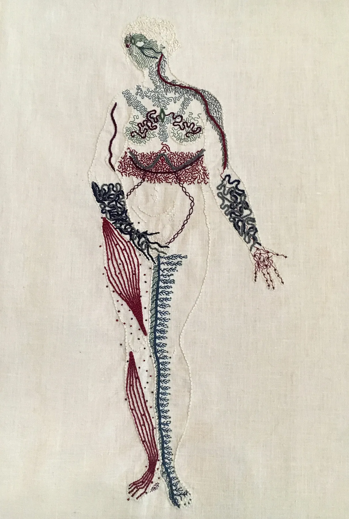 An outline of a naked woman is embroidered on linen in the same bone white colour as the linen. She stands legs together, her right hand covering her groin, her left hand, palm up, extended slightly to her side. She looks to the right. Her entire body except for her belly is covered in intricate markings representing different neurological sensations. Her face is a mask of green lines, feathery grey lines cover her shoulders and chest. There is a thick band of intricate burgundy stitching around her waist. Her forearms and hands are covered in thick blue undulant lines. Her right leg has bands of burgundy along the muscles, with small dots around them. Her inner left leg has a thick line of blue running up it, with thin branches spreading towards her outer leg.