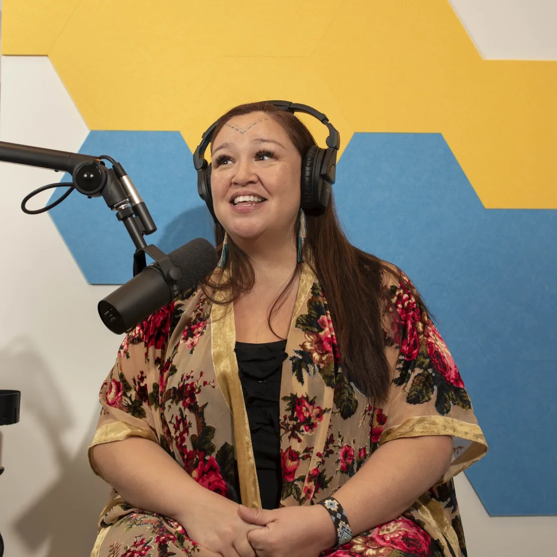 Indigenous person in a floral print top speaks on a podcast microphone in front of a yellow and blue tiled wall