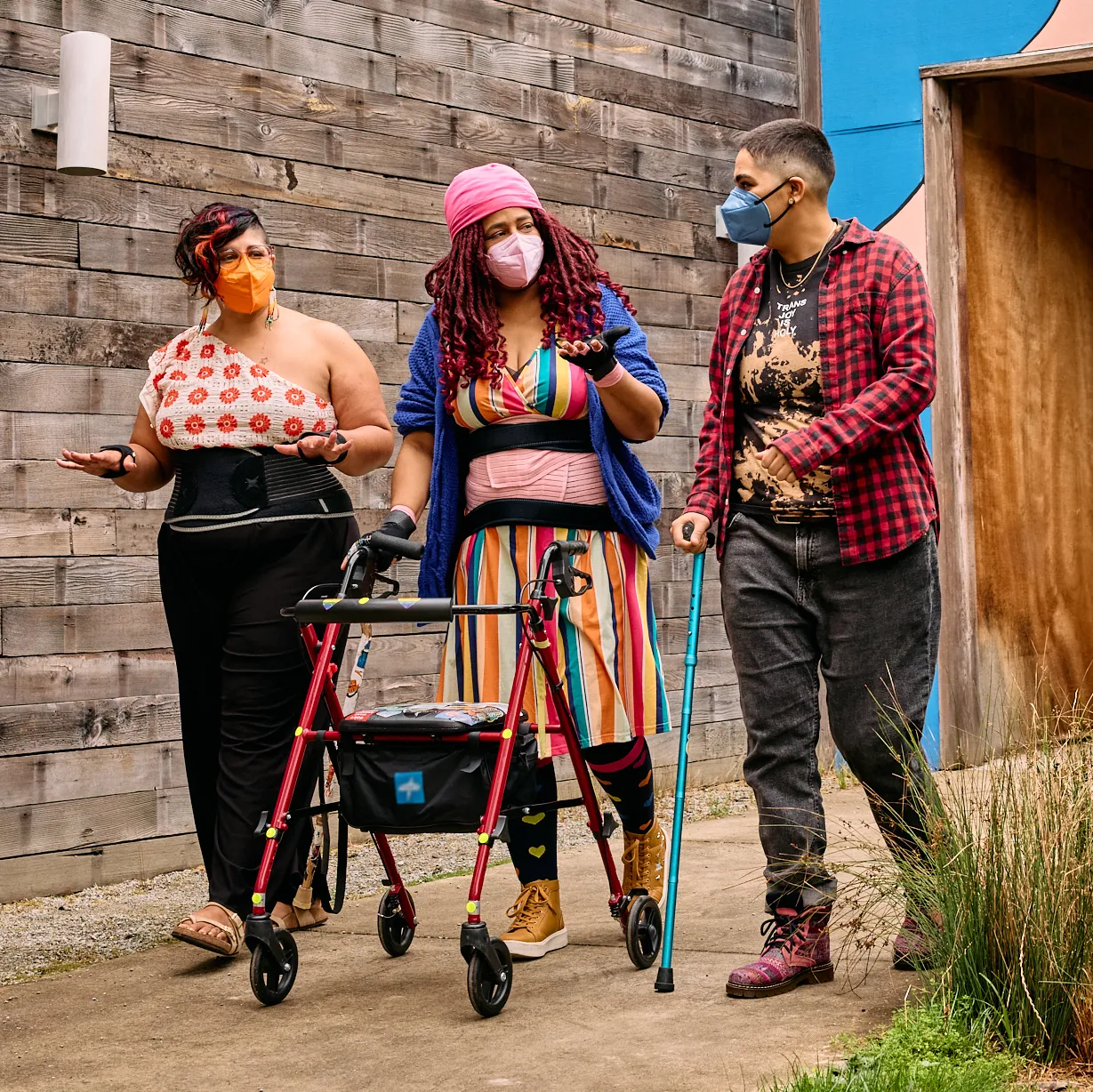 Three masked disabled people of color chat while strolling through an outdoor courtyard. On the left, an Indigenous woman wears glasses, wrist support braces, and waist trainers paired with a off-shoulder top and wide leg pants. In the middle, a Black non-binary woman walks with a rollator and wears waist trainers with ice pack inserts over their rainbow striped dress. On the right, a Latinx non-binary person walks while using a teal functional grip cane.