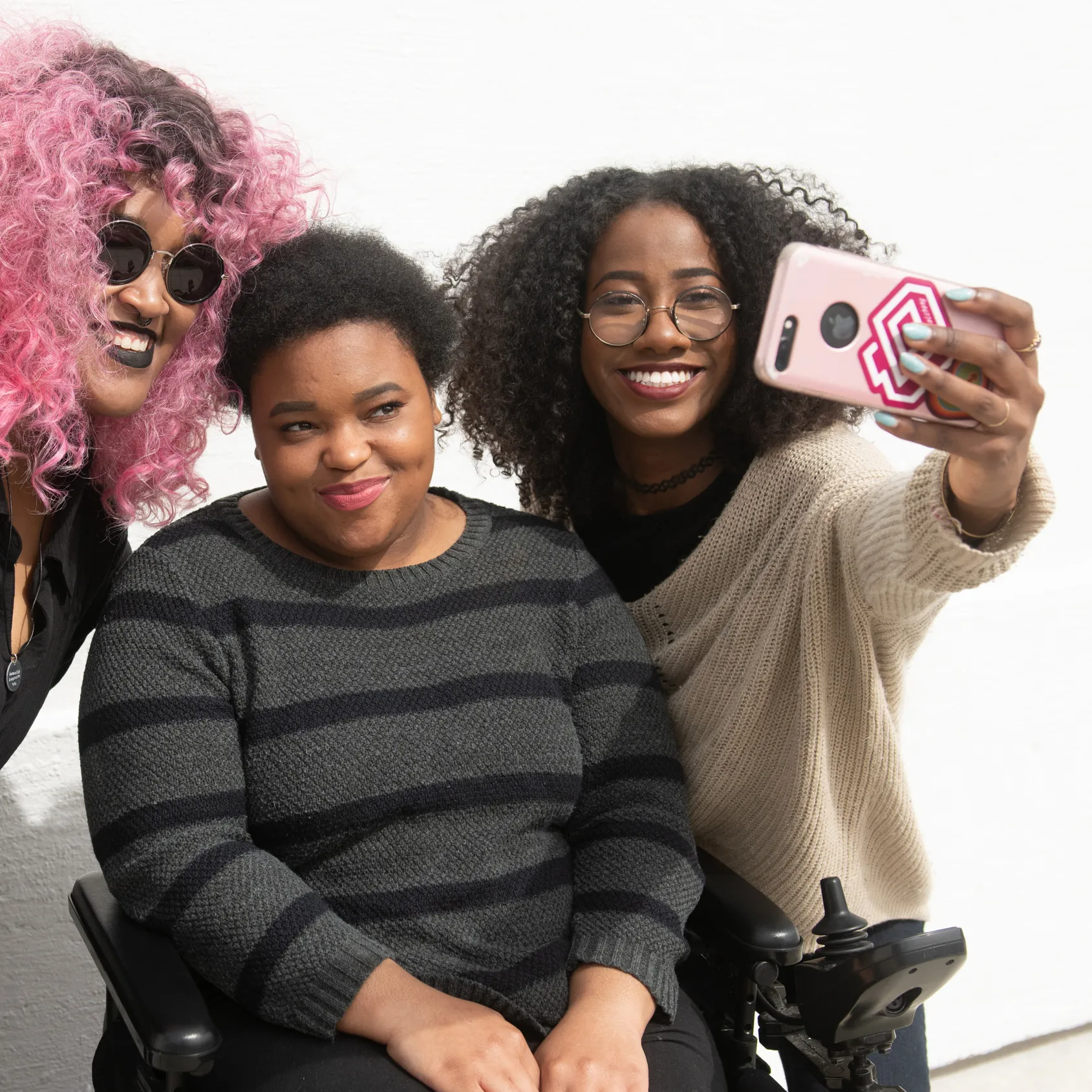 A tight crop of three Black and disabled friends, including one seated in a power wheelchair, taking a mobile phone selfie together.