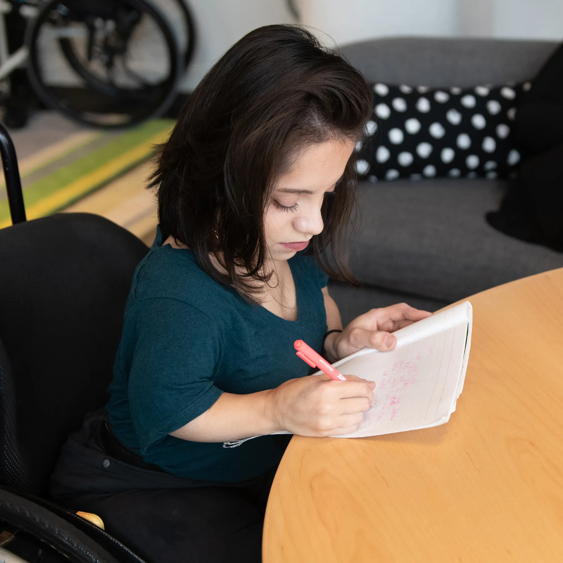 A South Asian person in her wheelchair takes notes by hand during a meeting.