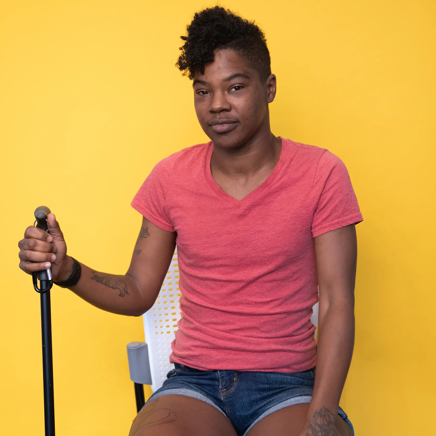 A Black non-binary person with a short mohawk and tattoos sits on a chair in front of a yellow backdrop while holding a black cane. They have a neutral expression and are wearing a salmon T-shirt with jean shorts.