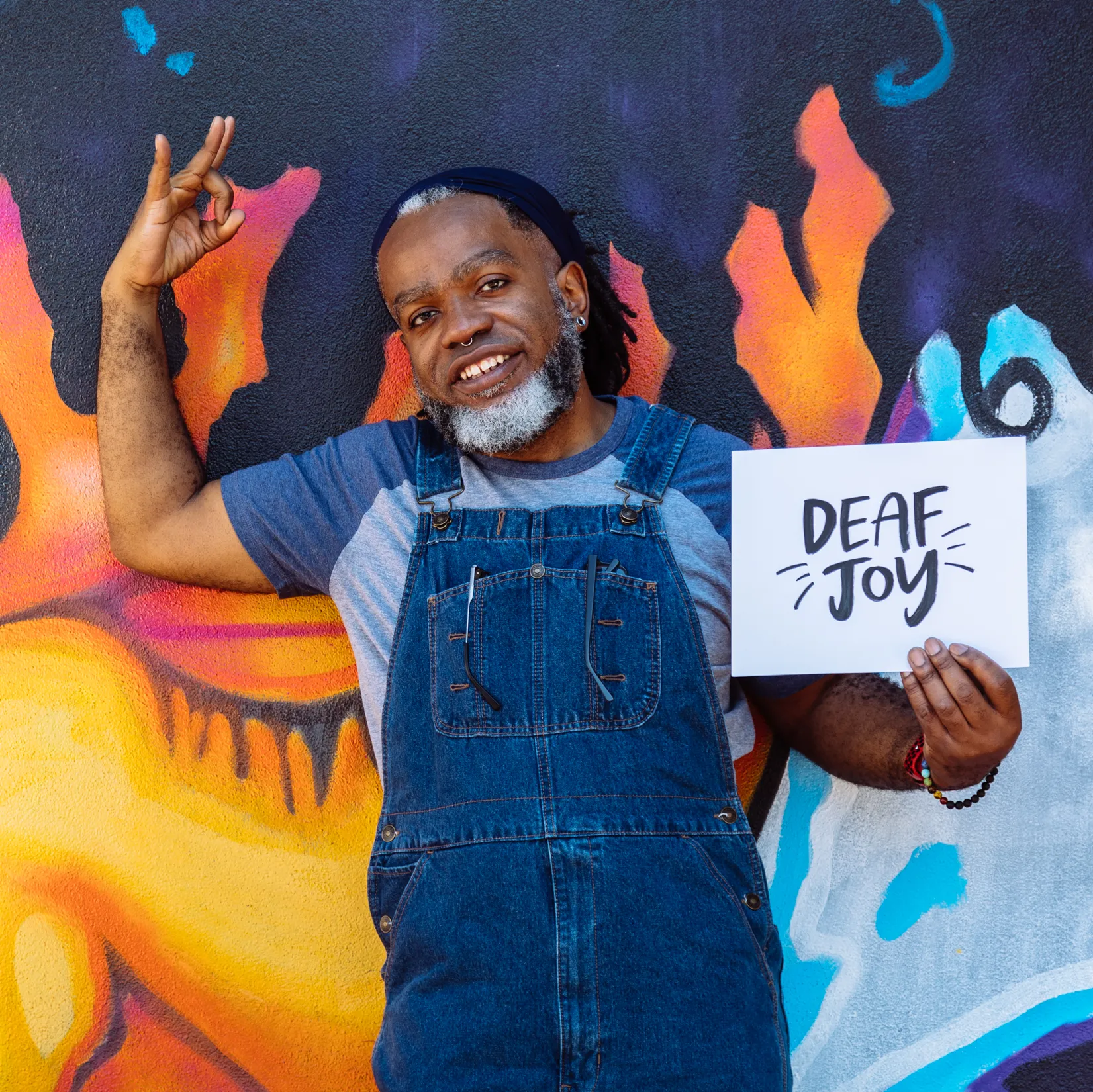A Deaf Black man smiles and signs in American Sign Language while holding a “Deaf joy” print. The man is wearing glasses and denim overalls and has a black and white beard, septum piercing, and hair pulled back by a bandana. The background features a vibrant mural with a Black woman’s face in close-up.