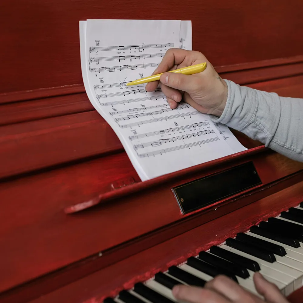 At a red piano, a composer has one hand on the piano keys and the other making notations in pencil on a page of sheet music