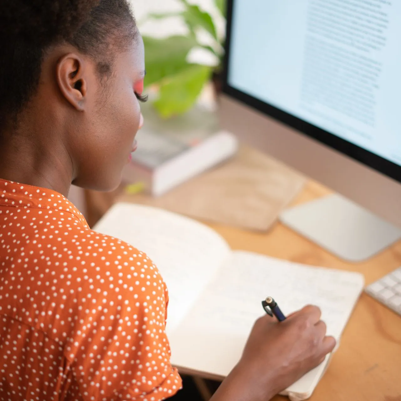 A Black person in an orange shirt is writing in a notebook at a desk.