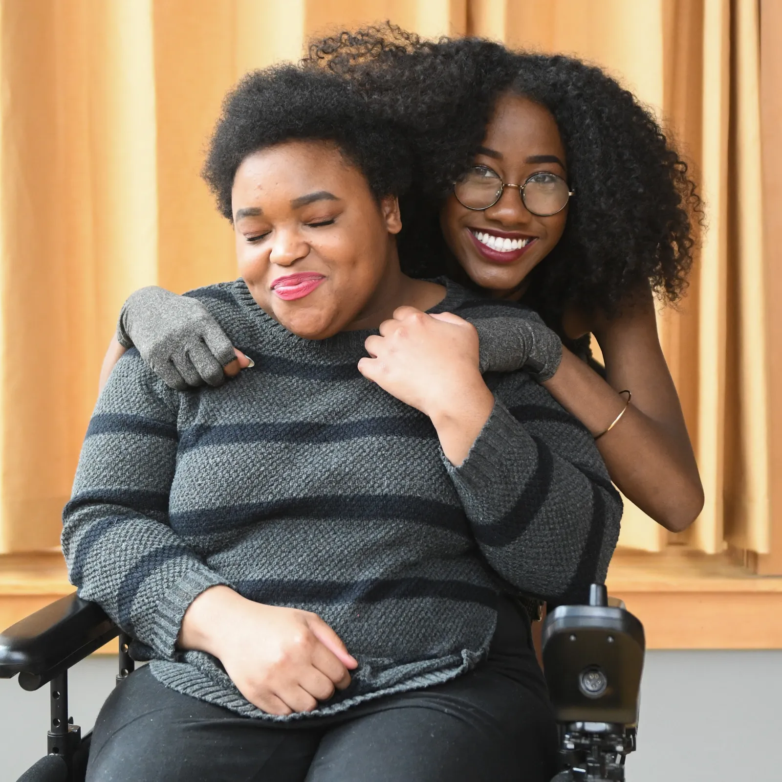 A Black non-binary person in a black and pink power wheelchair has their eyes closed and is smiling while being hugged from behind by a Black femme wearing compression gloves.
