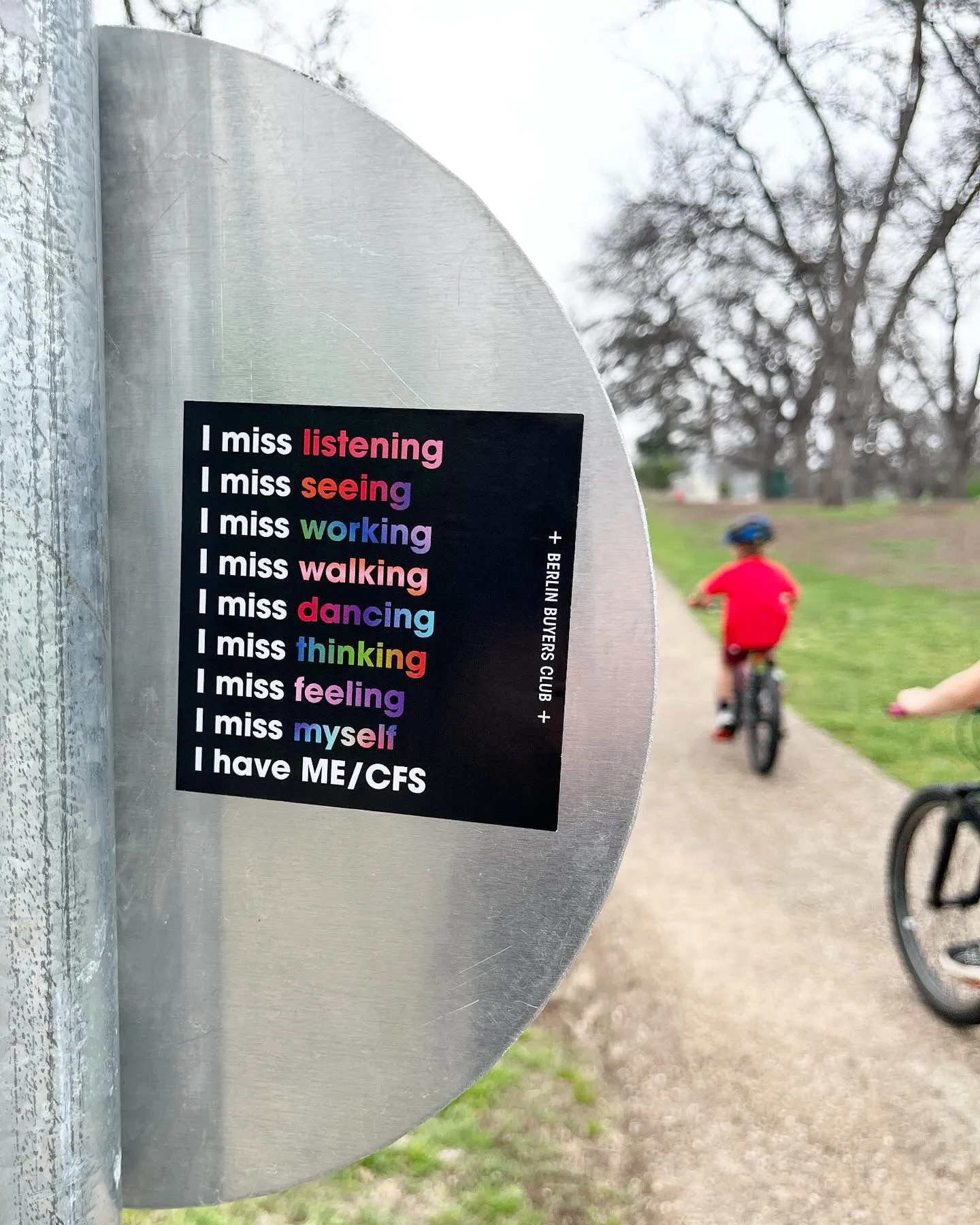 Photo of an outside scene. A bike path is in the background next to some grass with trees in the distance. People are riding towards the horizon. In focus, there is a sticker on the back of a sign; it's black, and there is text in white that says: "I miss listening, I miss seeing, I miss working, I miss walking, I miss dancing, I miss thinking, I miss feeling, I miss myself, I have ME/CFS" the words listening, seeing, working, walking, dancing, thinking, feeling and myself are in multicoloured gradients.