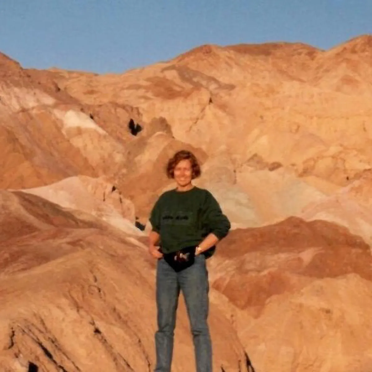 Archival photo of a young Eileen Stephens before she got sick. She is a brown-haired white woman wearing a dark sweatshirt, jeans and belt bag, standing confidently with her hands on her hips in front of a mountainous desert landscape.