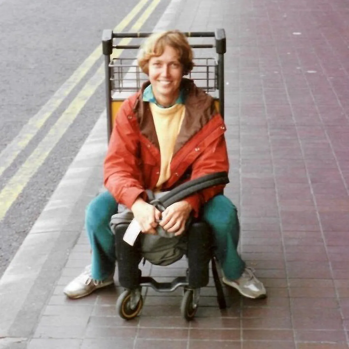 Archival photo of a young Eileen Stephens before she got sick. She is a young white woman with short light brown hair, dressed in casual clothes including a red jacket, sitting on a luggage cart, grinning.