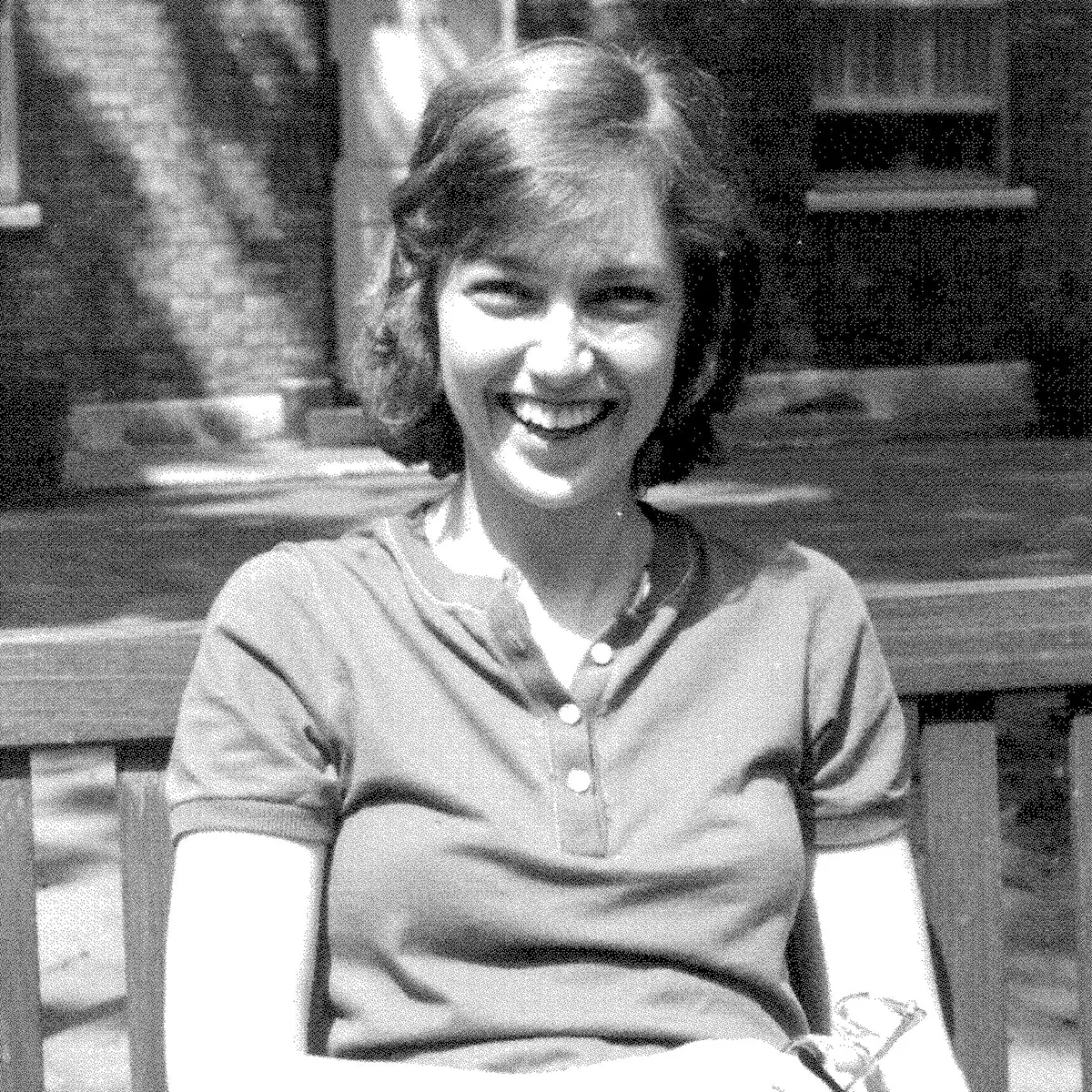 Archival black and white photo of a young Eileen Stephens before she got sick. She has short hair and is wearing a short-sleeved collarless polo shirt and jeans. She is sitting on a bench outdoors, grinning.