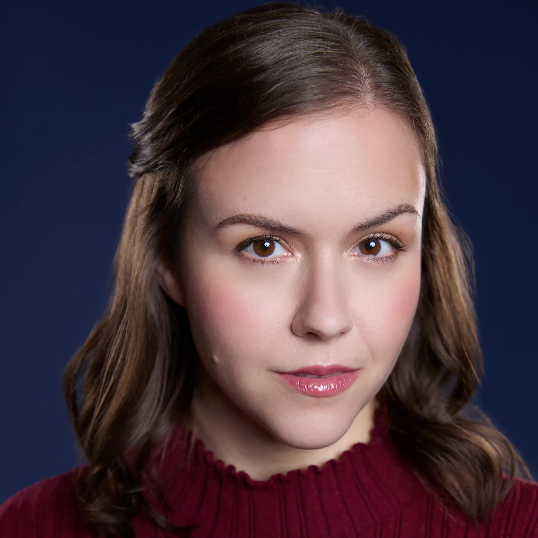 Headshot of Robin, a white femme person with shoulder length brown hair and brown eyes. She is wearing a wine red sweater, and the background is dark blue.