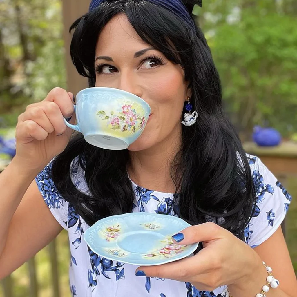 Outdoors on a pretty patio, Christina Baltais sips tea from a blue foral teacup and saucer. Christina is a white woman with dark brown hair in a retro bouffant hairstyle with blue headband. She wears a blue and white floral dress and matching teapot jewelry.