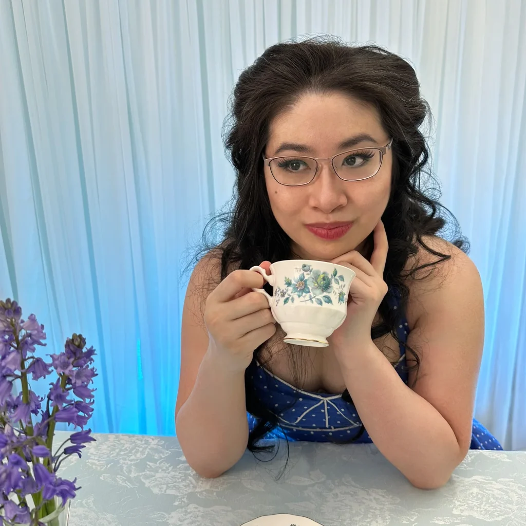 Stephanie is seated at a table with a light blue tablecloth, in front of white curtains lit in blue, holding a blue and white porcelain teacup. She's a Chinese Canadian woman with black wavy hair and glasses, wearing a blue and white polkadot dress. On the table in front of her is a teacup saucer and a vase of bluebells.