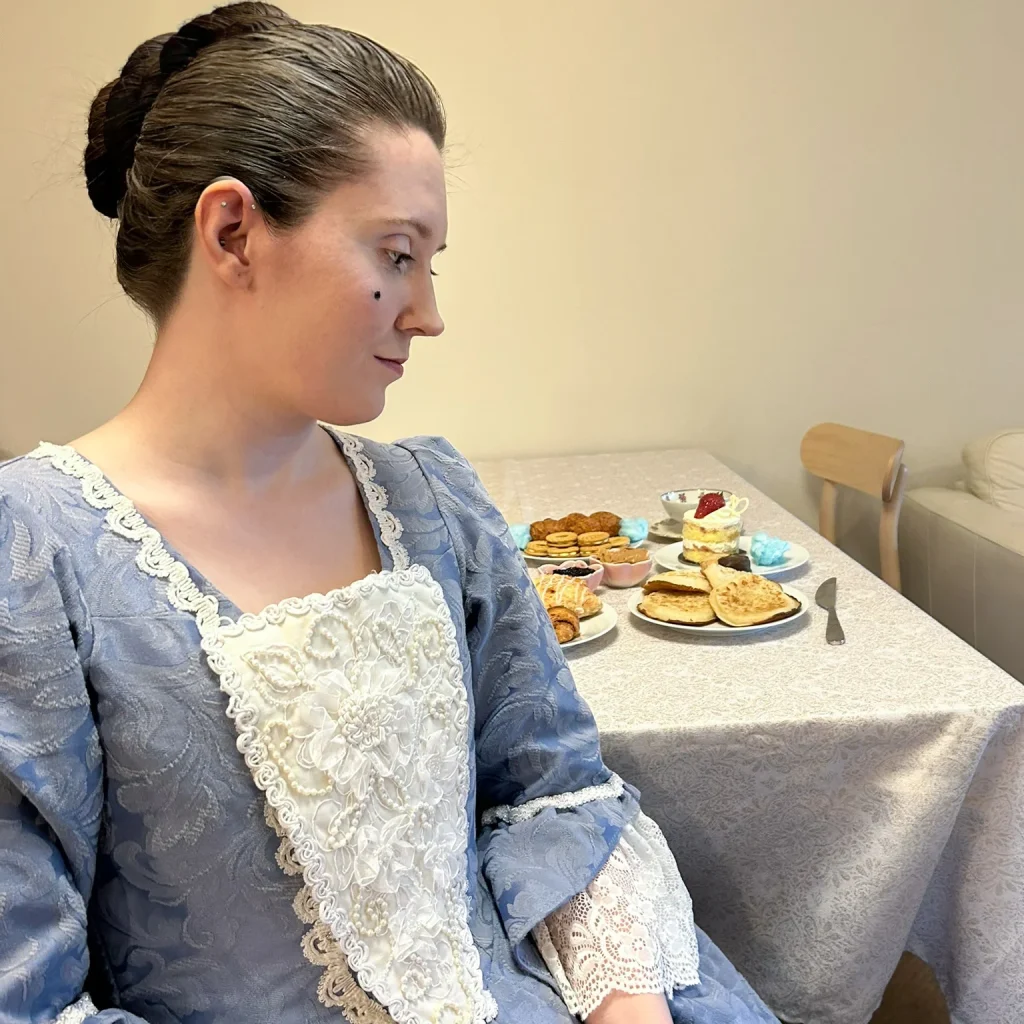 Christie, a white woman with brown hair in an updo, sits in profile wearing a sky blue Georgian gown with a pearl-encrusted white stomacher and lace cuffs. Behind her is a table spread with dessert, pastries, and puffs of blue cotton candy.