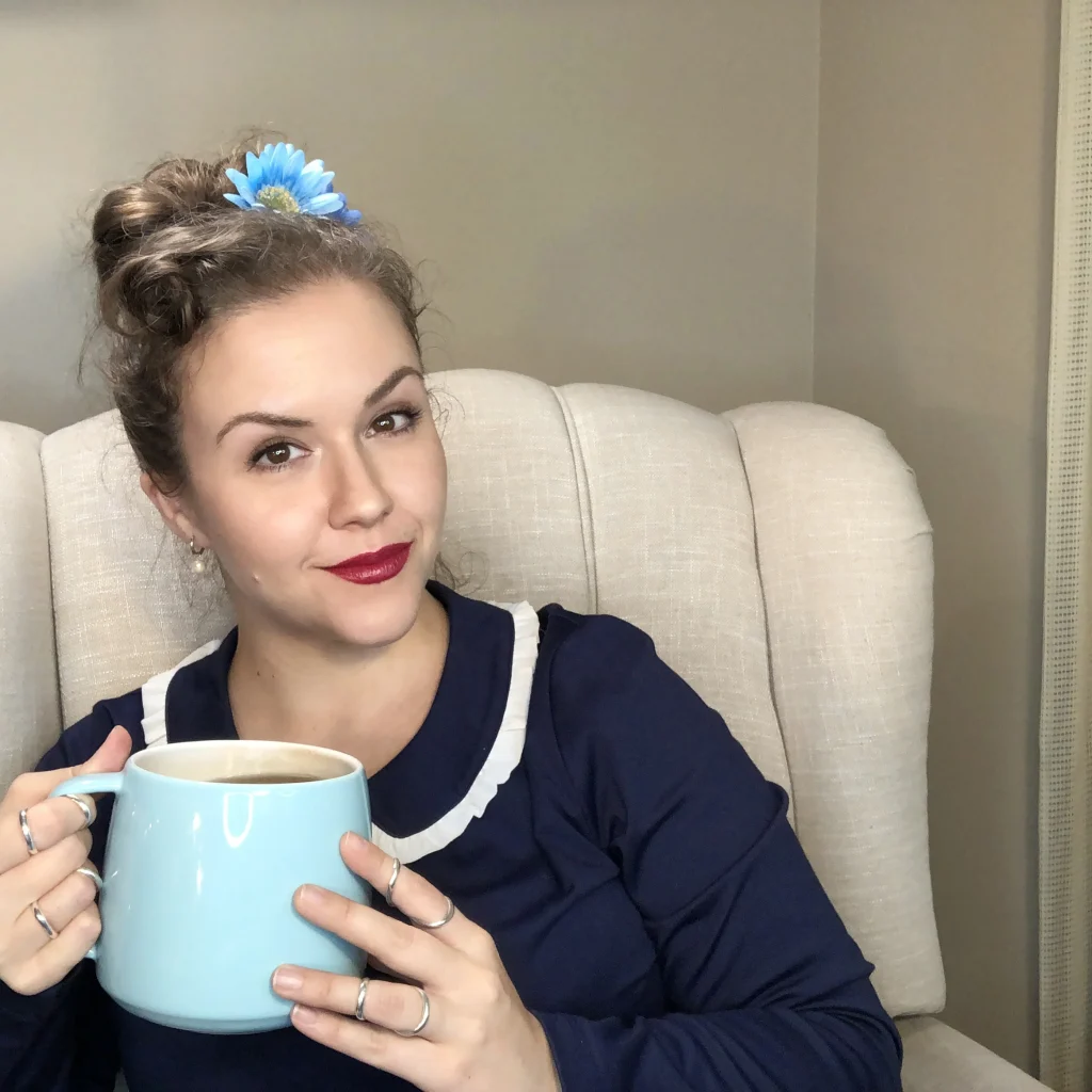 Robin sits in a beige armchair, holding an oversized cup of blue tea. Robin is a white femme person with blonde hair in an updo with a blue flower, wearing a navy blue dress with Peter Pan collar, red lipstick, and silver ring splints.